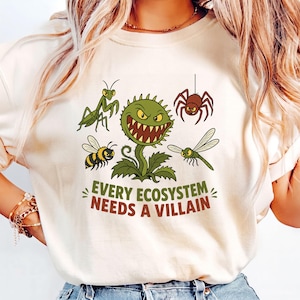 May include: A cream-colored t-shirt featuring a graphic print of a central green Venus flytrap surrounded by a praying mantis, a spider, a bee, and a dragonfly. Below the insects, the text reads, "EVERY ECOSYSTEM NEEDS A VILLAIN" in green and brown.