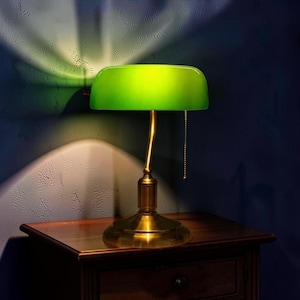 May include: A desk lamp featuring a vibrant green glass shade and a metallic brass-toned base. The lamp has a curved stem and a pull chain switch, emitting a bright, focused light that casts a distinct, fan-shaped glow against the wall behind it.