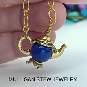 May include: A gold-colored teapot pendant with a deep blue gemstone body, suspended from a gold chain. The teapot features intricate details, including a handle, spout, and lid. The text "MULLIGAN STEW JEWELRY" is displayed below the pendant.