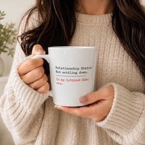 Peut inclure: Un mug en céramique blanche avec un texte noir indiquant Relationship Status: Not settling down. Une ligne horizontale noire sépare ce texte de la phrase en rouge In my Littlest Hobo era. Le mug possède une anse incurvée sur le côté gauche.