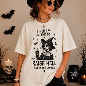 May include: An off-white short-sleeve t-shirt featuring a black graphic print of a witch wearing a hat and sunglasses, holding a coffee cup. The text reads: I FULLY INTEND TO RAISE HELL AND DRINK COFFEE, surrounded by small stars, bats, and a crescent moon.