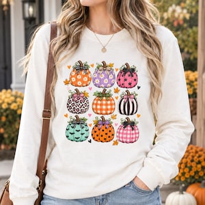 Puede incluir: Una sudadera de manga larga color crema con un estampado gráfico de nueve calabazas con diversos patrones, incluyendo flores, leopardo, cuadros, rayas y lunares, cada una rematada con un pequeño lazo y rodeada de pequeñas hojas.