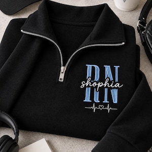 May include: A black quarter-zip sweatshirt featuring embroidered text in light blue and white. The embroidery displays the letters RN, the name "shophia" in script, and another RN below, finished with a white heartbeat line and a small heart symbol.