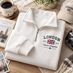 Puede incluir: Una sudadera blanca con cremallera de un cuarto y un gráfico bordado en azul marino en el pecho que dice LONDON EST. 1947 United Kingdom, acompañado de una pequeña bandera Union Jack y un icono de corona. Tiene cuello alto y puños acanalados.