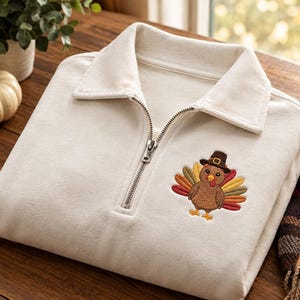 May include: A cream-colored quarter-zip sweatshirt featuring a folded collar and a silver-toned metal zipper. An embroidered graphic of a brown turkey wearing a dark brown pilgrim hat with a yellow buckle is positioned on the left chest area.