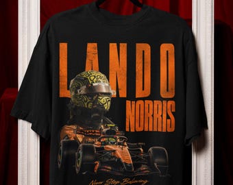 May include: A black t-shirt featuring a large orange graphic of Lando Norris in a racing car with a yellow helmet. The text reads LANDO NORRIS in bold orange letters and Never Stop Believing in a smaller script font below the car graphic.