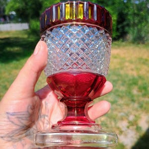 May include: A clear and ruby red glass goblet featuring a diamond-cut pattern on the bowl, a smooth red stem, and a square base. The base is etched with the text "L. Barbero 1980". The rim of the goblet is a deep red with vertical fluting.