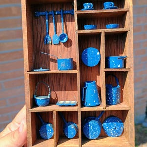 May include: A miniature wooden display shelf containing a collection of blue and white speckled enamel kitchenware. The set includes tiny pots, pans, cups, a pitcher, a kettle, and hanging utensils, all arranged within the divided wooden compartments.