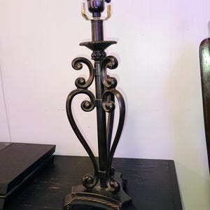 May include: A dark bronze-coloured metal table lamp base featuring a central vertical rod surrounded by decorative curved scrollwork. The lamp has a tripod-style base with rope-like detailing and a standard light bulb socket at the top.