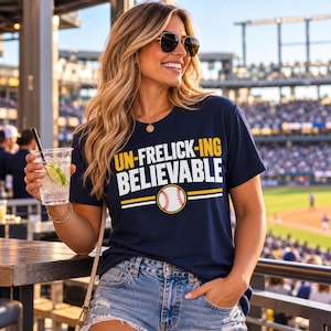 May include: A navy blue short-sleeved t-shirt featuring the yellow and white text UN-FRELICK-ING BELIEVABLE with a graphic of a baseball in the center. The shirt has a crew neckline and is made of a solid dark blue fabric.