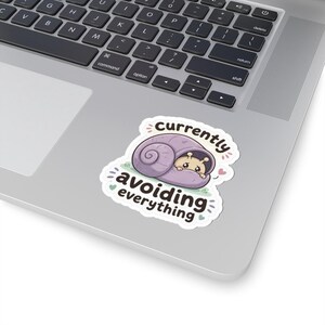 May include: A die-cut sticker featuring a cartoon snail with a light purple shell and a tan body peeking out. The text above and below the snail reads "Currently avoiding everything" in dark brown letters, accented by small green hearts and pink dots.