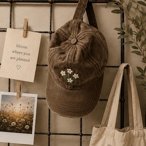 May include: A brown baseball cap with a faded, distressed texture featuring an embroidered design of three small white flowers with yellow centers on the front panel. The cap has a curved brim and a matching brown adjustable strap at the back.