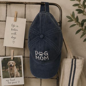 May include: A dark blue baseball cap with a washed-out finish featuring white embroidered text that reads DOG MOM with a paw print icon above the word DOG. The cap has a structured crown, a curved brim, and a fabric strap at the top.