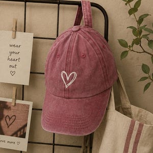 Può includere: Un cappellino da baseball color rosa polvere con il contorno di un cuore ricamato in bianco sul pannello frontale. Il cappellino presenta una texture in tessuto lavato e usurato, una visiera curva e un cinturino in tessuto con fibbia.