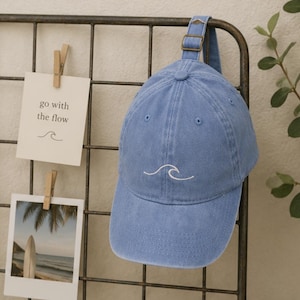Può includere: Un berretto da baseball azzurro con un piccolo disegno a onda ricamato in bianco sul pannello frontale. Il berretto include un cinturino regolabile con fibbia in metallo sul retro per una vestibilità sicura. Il tessuto ha un aspetto slavato.