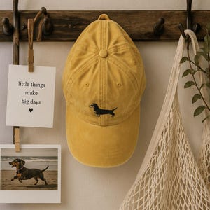 Pode incluir: Um boné de beisebol amarelo mostarda com um pequeno cão dachshund preto e castanho bordado no painel frontal. O boné tem uma coroa estruturada com um botão no topo, ilhós costurados e uma aba curva num design clássico de seis painéis.