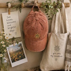 May include: A rust-coloured baseball cap with an embroidered yellow sun design, paired with a beige canvas tote bag featuring a black sun graphic and the text 'you are my sunshine' with a heart symbol, hanging on a wooden wall rack.