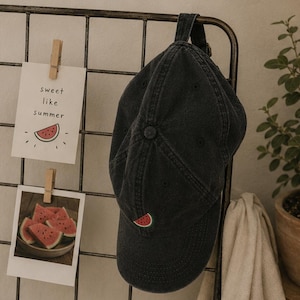 May include: A charcoal grey baseball cap featuring a small embroidered watermelon slice on the front panel. The cap has a six-panel construction with a button on top and an adjustable strap at the back. The fabric has a washed, faded appearance.