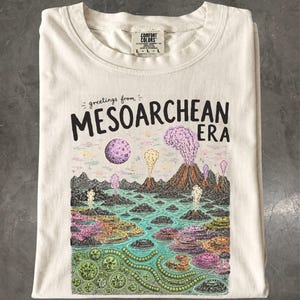 Peut inclure: Un t-shirt couleur crème avec une impression graphique portant le texte "greetings from MESOARCHEAN ERA". L'illustration représente un paysage préhistorique avec des montagnes volcaniques, des cratères en éruption, une lune violette et des formes de vie microbiennes colorées.