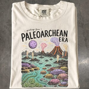 Pode incluir: Uma t-shirt de cor creme com uma impressão gráfica com o texto "greetings from PALEOARCHEAN ERA". A ilustração retrata uma paisagem vulcânica pré-histórica com formações geológicas roxas, cor-de-rosa e amarelas, uma lua e água.