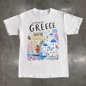 Peut inclure: Un t-shirt blanc à manches courtes avec une impression graphique colorée portant le texte Greetings from GREECE. L'illustration représente le Parthénon, des bâtiments à dômes bleus, un drapeau grec, un bateau, un voilier, une amphore et des fleurs.