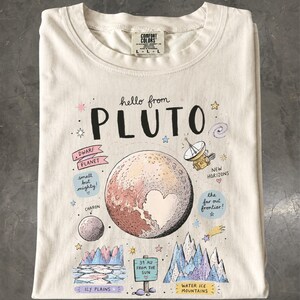 Pode incluir: Uma t-shirt de cor creme com um gráfico de Plutão que apresenta uma forma de coração na sua superfície. O design inclui o texto hello from PLUTO, DWARF PLANET, small but mighty, NEW HORIZONS, the far out frontier, CHARON, 39 AU FROM THE SUN, ICY PLAINS e WATER ICE MOUNTAINS.