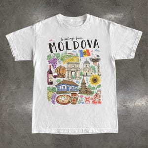 May include: A white short-sleeve t-shirt featuring a colourful graphic print with the text "Greetings from MOLDOVA". The illustration includes a wine bottle, grapes, a barrel, a triumphal arch, a cathedral, a stork, a sunflower, and traditional food.