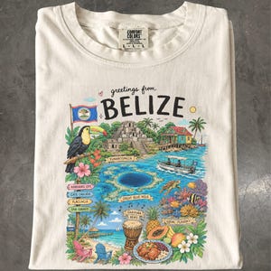 Pode incluir: Uma t-shirt cor de creme com uma impressão gráfica colorida com o texto "greetings from BELIZE". O design inclui um tucano, a bandeira do Belize, as ruínas de Xunantunich, o Great Blue Hole, um barco, um tambor e frutas tropicais.