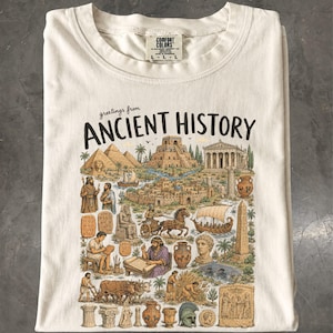 May include: A cream-coloured t-shirt featuring a graphic print with the text "greetings from ANCIENT HISTORY". The illustration depicts various historical landmarks, figures, and artefacts including pyramids, a chariot, and classical pottery.