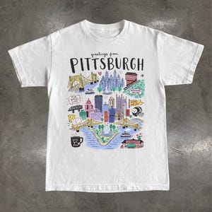 May include: A white short-sleeved t-shirt featuring a colourful hand-drawn illustration of the Pittsburgh skyline. The graphic includes the text "greetings from PITTSBURGH" and depicts iconic landmarks, bridges, a football helmet, and a coffee mug.