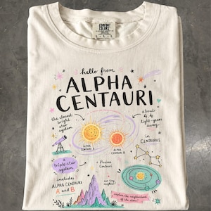 May include: A cream-colored t-shirt featuring a graphic print about the Alpha Centauri star system. The design includes text, illustrations of stars, a telescope, a mountain range, and planetary orbits, with labels detailing facts about the system.
