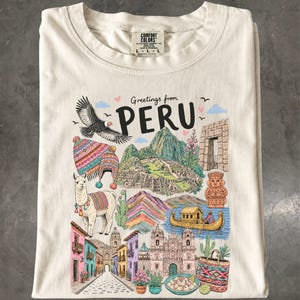Op de afbeelding: Een crèmekleurig t-shirt met een kleurrijke grafische print met de tekst "Greetings from PERU". De illustratie bevat een condor, een lama, Machu Picchu, een rieten boot, een stenen deuropening, een standbeeld en levendige straatbeelden.