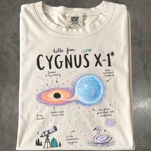 May include: A cream-colored t-shirt featuring a graphic print titled hello from CYGNUS X-1. The design includes a black hole, a blue supergiant star, a telescope, and a swan, with handwritten labels describing the astronomical features.