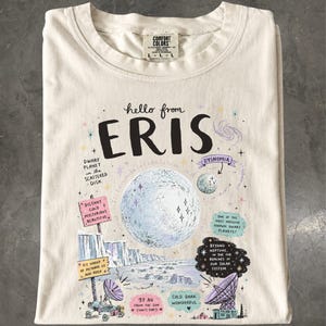 Może przedstawiać: Kremowy t-shirt z grafiką przedstawiającą planetę karłowatą Eris. Wzór zawiera dużą centralną planetę, mniejszy księżyc o nazwie Dysnomia, ilustracje o tematyce kosmicznej oraz tekst z faktami dotyczącymi odległości i składu tej planety.