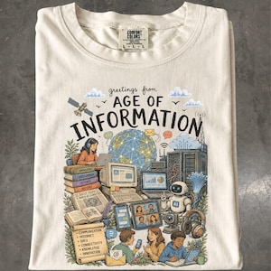 Può includere: Una t-shirt color crema con una stampa grafica che riporta il testo "greetings from AGE OF INFORMATION". L'illustrazione raffigura un globo, computer, un robot, libri e persone che utilizzano la tecnologia, con un elenco di termini come comunicazione e dati.