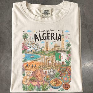 May include: A cream-colored t-shirt featuring a colorful graphic print with the text "Greetings from ALGERIA". The illustration depicts a coastal city, a mosque, the Algerian flag, desert dunes with camels, a fennec fox, and traditional food.