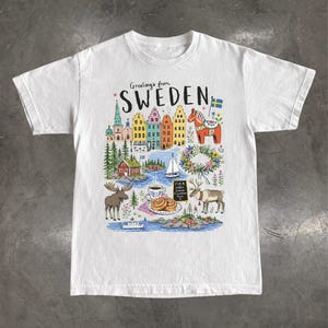May include: A white short-sleeve t-shirt featuring a colourful graphic illustration with the text "Greetings from SWEDEN". The design includes Swedish buildings, a Dala horse, a sailboat, a moose, a reindeer, a floral wreath, and cinnamon rolls.