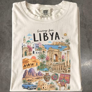 May include: A cream-colored t-shirt featuring a graphic print with the text Greetings from LIBYA. The illustration includes a coastal scene with a sailboat, ancient stone arches, desert dunes with camels, a blue car, and traditional pottery.