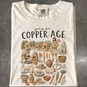 Könnte beinhalten: Ein cremefarbenes T-Shirt mit einem Grafikdruck und dem Text "greetings from COPPER AGE". Die Illustration zeigt eine prähistorische Dorfszene mit Menschen, Kupferwerkzeugen, Töpferwaren, einer Ziege, Weizen und steinernen Strukturen in Erdtönen.