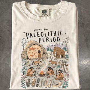 May include: An off-white t-shirt featuring a graphic print with the text "greetings from PALEOLITHIC PERIOD". The illustration depicts a cave scene with prehistoric humans, a mammoth, deer, a campfire, stone tools, and cave wall paintings.