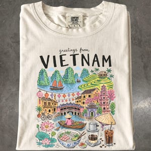 Op de afbeelding: Een crèmekleurig t-shirt met een kleurrijke grafische print met de tekst "greetings from VIETNAM". De illustratie toont een landschap met kalkstenen rotsen, rijstterrassen, een brug, een boot, lotusbloemen, een kom pho en koffie.