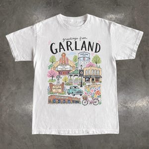 May include: A white short-sleeve t-shirt featuring a colorful graphic illustration with the text "greetings from GARLAND". The design depicts various landmarks, a water tower, a vintage truck, a bicycle, and trees in a detailed, hand-drawn style.