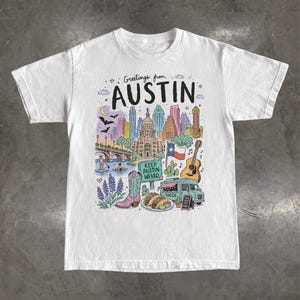 May include: A white short-sleeve t-shirt featuring a colourful graphic illustration of the Austin skyline, a cowboy boot, a guitar, tacos, a food truck, and the text "Greetings from AUSTIN" and "KEEP AUSTIN WEIRD" printed on the front centre.