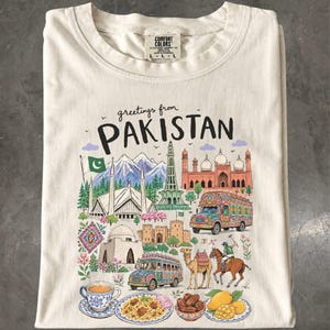 May include: A cream-colored t-shirt featuring the text greetings from PAKISTAN above a colorful illustration of landmarks, a decorated truck, a camel, a polo player, and traditional food items including a cup of tea and a plate of biryani.