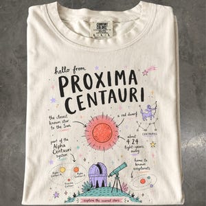 Puede incluir: Una camiseta de color crema con un estampado gráfico que incluye el texto "hello from PROXIMA CENTAURI". El diseño muestra una estrella enana roja, un telescopio, una constelación y diagramas del sistema Alfa Centauri con varias etiquetas.