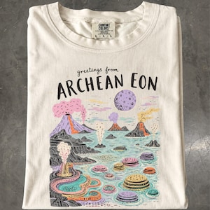 Pode incluir: Uma t-shirt em tom off-white com uma estampa gráfica contendo o texto "greetings from ARCHEAN EON". A ilustração retrata uma paisagem pré-histórica com vários vulcões em erupção, uma lua roxa grande e formações terrestres circulares coloridas.