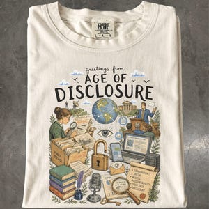 May include: A cream-colored t-shirt featuring a graphic print with the text "greetings from AGE OF DISCLOSURE". The illustration includes a globe, a person at a podium, a magnifying glass, a padlock, a microphone, books, and documents.