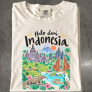 Puede incluir: Una camiseta de color crema con un estampado gráfico colorido con el texto "Halo dari Indonesia". La ilustración muestra monumentos indonesios, terrazas de arroz, un dragón de Komodo, una cacatúa y un barco en un estilo multicolor.