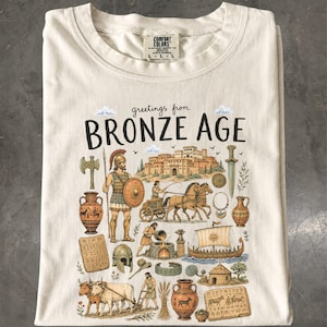 May include: A cream-coloured t-shirt featuring a graphic print with the text "greetings from BRONZE AGE". The illustration depicts historical scenes including a warrior, a chariot, a ship, pottery, livestock, and ancient architecture in earth tones.