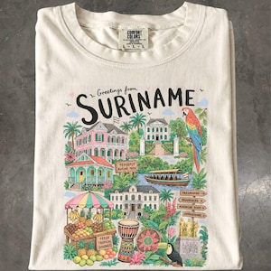 May include: An off-white crew neck t-shirt featuring a colourful graphic print with the text Greetings from SURINAME. The illustration depicts colonial buildings, a parrot, a fruit stand, a boat, a drum, and a signpost with various locations.